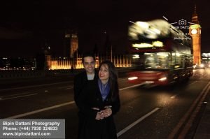 proposal photographer london with happy couple after proposal on Westminster Bridge. Houses of Parliament with Big Ben and the iconic London red bus zooming behind the couple